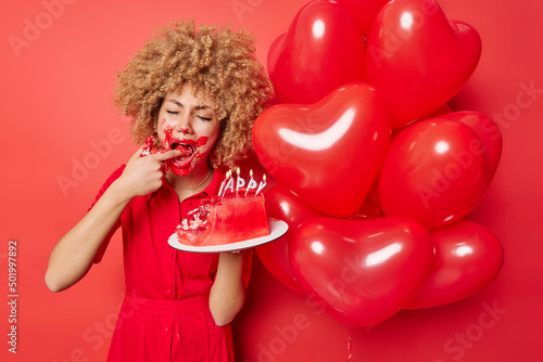 Birthday girl tastes cake with cream wears dress poses near bunch of inflated heart balloons has dirty mouth isolated over vivid red background. Upset curly woman sad to celebrate holiday alone