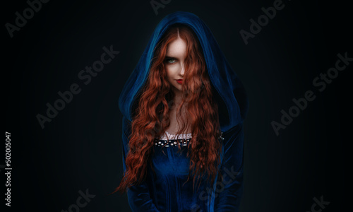 Portrait fantasy gothic red-haired woman witch. Vampire girl in blue medieval dress, vintage old historical style hood on head. Black background. Red lips, hair flying soar in wind. halloween costume