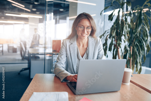 Smiling businesswoman with glasses working on laptop at her workplace in modern office. 