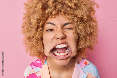 Headshot of curly haired young woman uses mouth expander special tool for whitetning shows white teeth winks eye dressed casually isolated over pink background. Stomatology and oral care concept