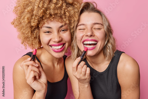 Happy beautiful women apply lipstick undergo beauty procedures stand next to each other dressed in black t shirts isolated over pink background. Two female friends prepare for party want to look good photo