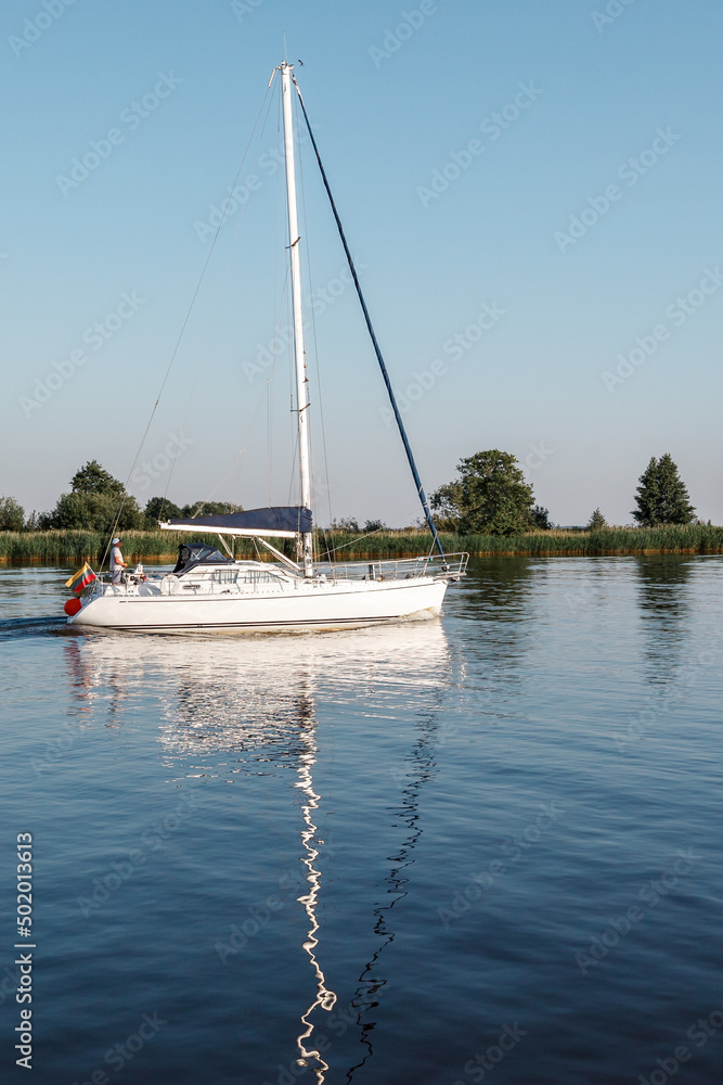 Obraz premium A white yacht with lowered sails and Lithuania flag in the lagoon. Quiet evening, well visible reflection of the ship's mast in the water. Vertical photo.