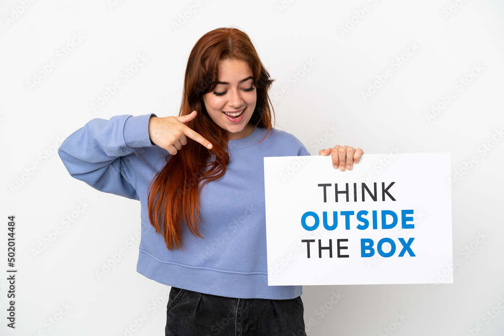Young redhead woman isolated on white background holding a placard with text Think Outside The Box and pointing it Young redhead woman isolated on white background holding a placard with text Think Outside The Box and pointing it