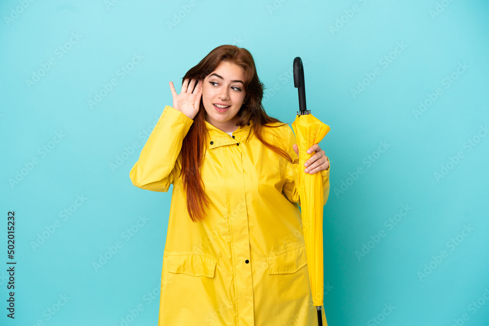 Redhead woman holding an umbrella isolated on blue background listening to something by putting hand on the ear Redhead woman holding an umbrella isolated on blue background listening to something by putting hand on the ear