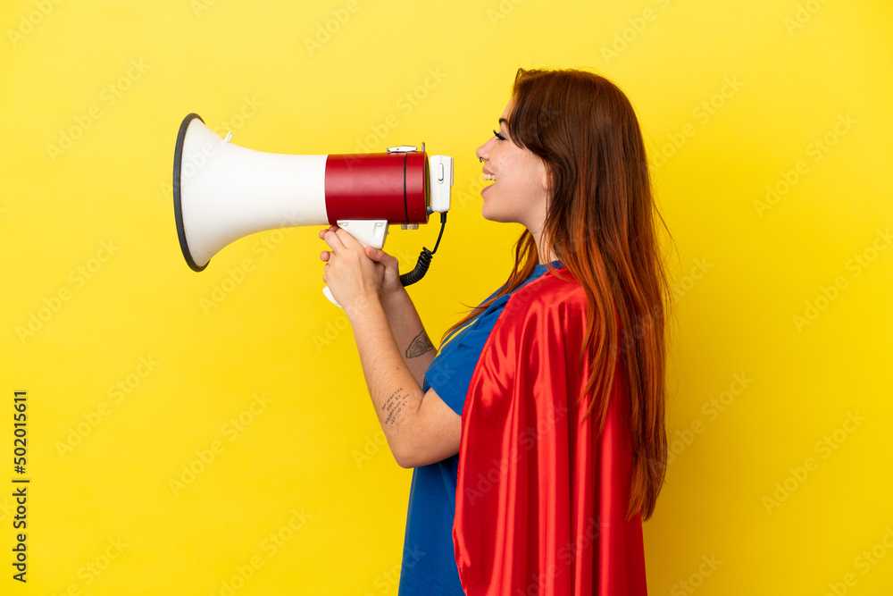 Super Hero redhead woman isolated on yellow background shouting through a megaphone Super Hero redhead woman isolated on yellow background shouting through a megaphone