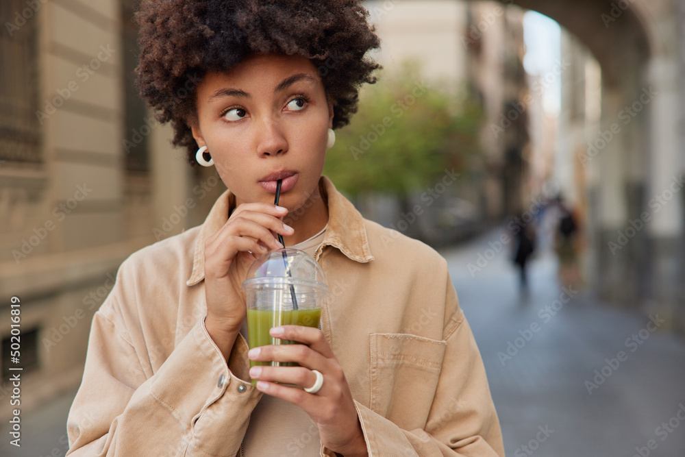Pretty thoughtful woman drinks detox beverage healthy organic green cocktail keeps to proper nutrition dressed in streetwear focused somewhere strolls in ancient city alone. Diet and lifestyle Pretty thoughtful woman drinks detox beverage healthy organic green cocktail keeps to proper nutrition dressed in streetwear focused somewhere strolls in ancient city alone. Diet and lifestyle