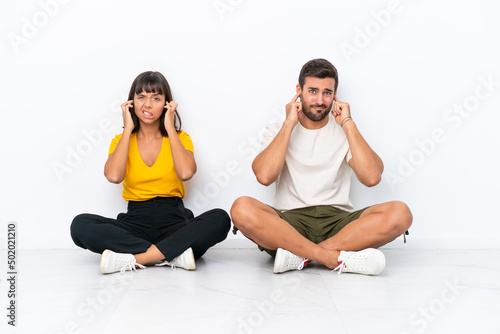 Young couple sitting on the floor isolated on white background covering both ears with hands