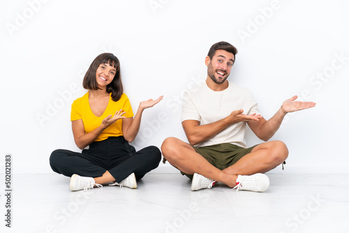 Young couple sitting on the floor isolated on white background extending hands to the side for inviting to come