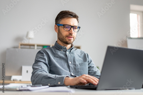 Focused mature businessman working with laptop computer at his desk in office, typing on pc keyboard, checking email