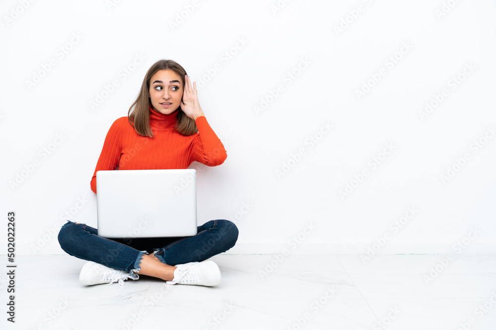 Young caucasian woman sitting on the floor with a laptop listening to something by putting hand on the ear Young caucasian woman sitting on the floor with a laptop listening to something by putting hand on the ear