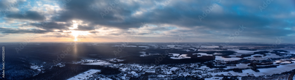Winter aerial wide panorama epic view on village, forest and fields covered in snow. Zmiyevsky region in Ukraine from drone. Sun shines through heavy sunset clouds