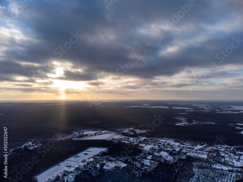 Winter aerial epic view on snowy village in forest. Zmiyevsky region in Ukraine from drone. Sun shines through heavy sunset clouds