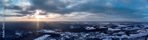 Winter aerial wide panorama epic view on village, forest and fields covered in snow. Zmiyevsky region in Ukraine from drone. Sun shines through heavy sunset clouds