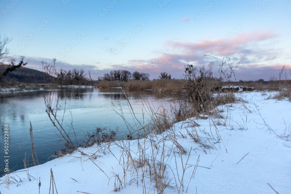 Fototapeta premium Cold river in winter, snowy coast close-up with scenic colorful clouds with reflection in water and dry reed grass. Zmiyevsky region on Siverskyi Donets River in Ukraine