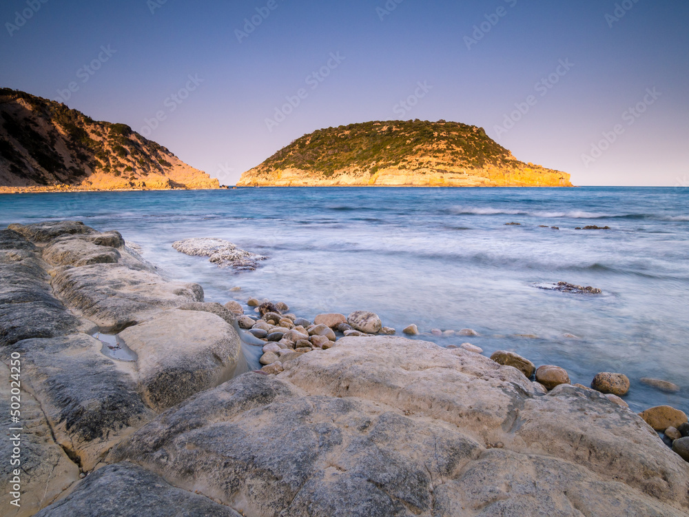 Paisaje costero con rocas en primer plano y una isla de fondo sobre cielo azul al atardecer