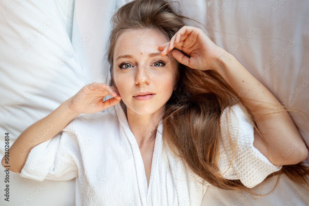 Fototapeta premium Portrait of young beautiful woman with long hair in white bathrobe on bed looking at camera in hotel or resort room. Cute attractive girl lying in bed on white sheets and pillows wear robe. Top view.