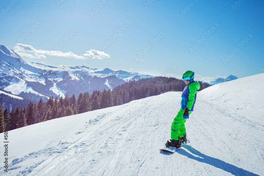 Boy in ski outfit stand on snowboard, sunny view over mountains