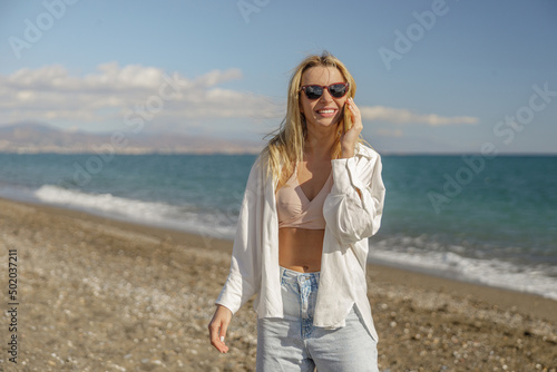 Excited blonde woman in sunglasses laughing while talking with someone over phone, standing on beach