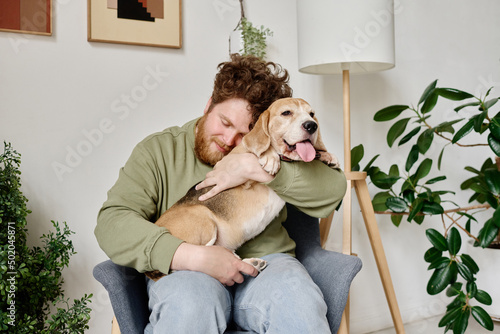 Young bearded man sitting on armchair in room with plants embracing his dog and relaxing photo