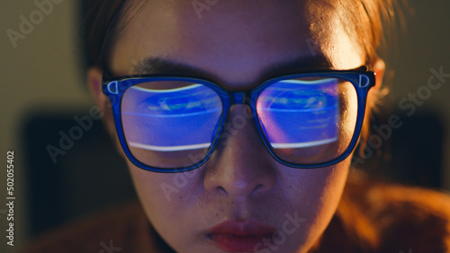 Close up of face of young Asian woman, developer programmer, software engineer, IT support, wearing glasses working hard at night overtime on computer to check coding in bugging system.