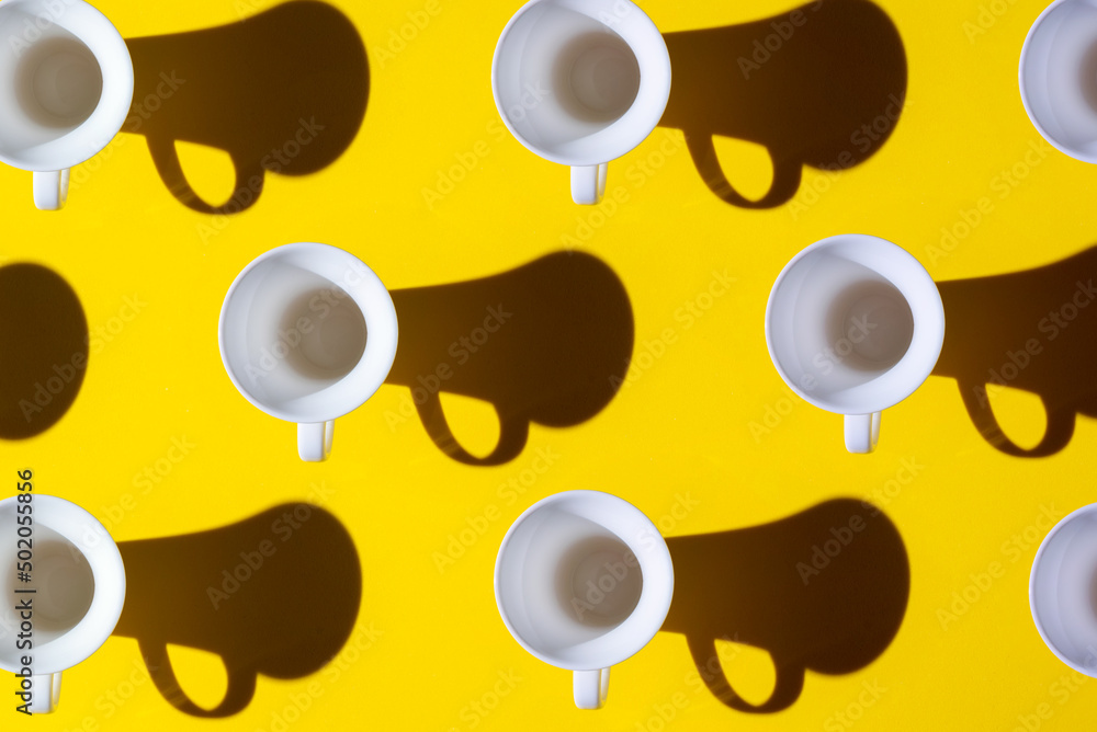 Pattern of coffee cups from a top view casting a hard shadow with the silhouette of another cup. Yellow background.