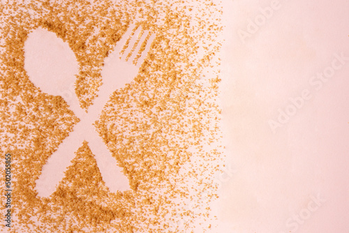 Crossed spoon and fork figure created in relief of muscovado sugar on pastel pink background. Copy space. photo