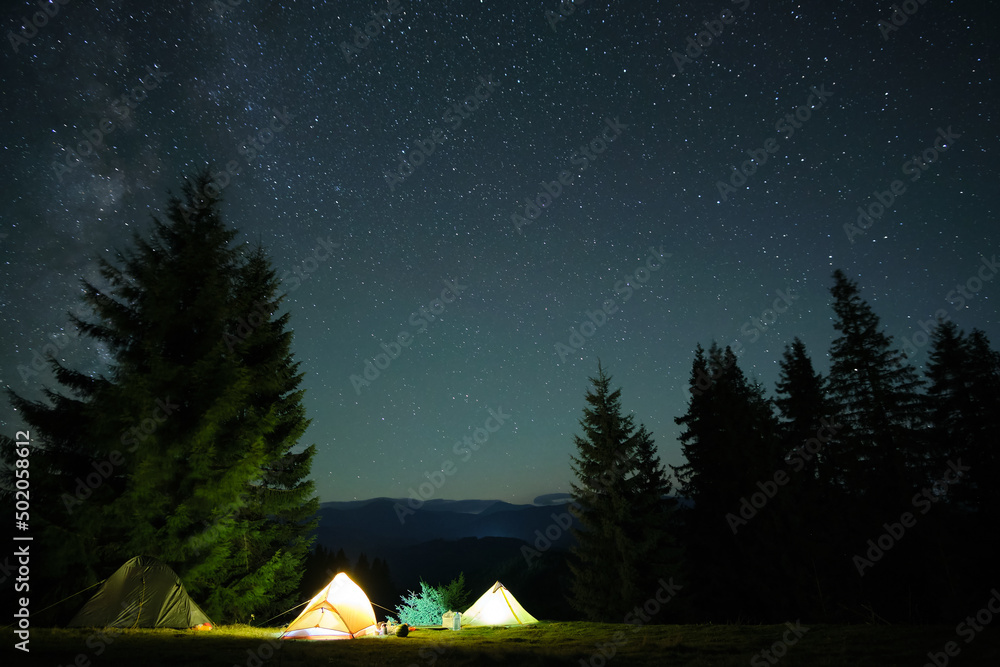 Bright illuminated tourist tents glowing on camping site in dark mountains under night sky with sparkling stars. Active lifestyle concept - obrazy, fototapety, plakaty 