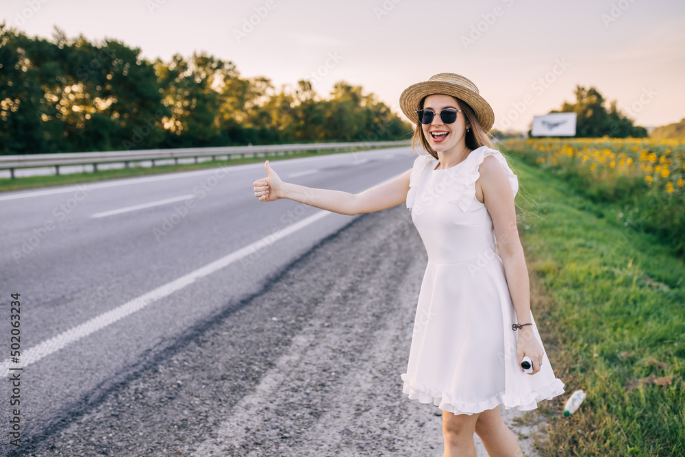 The girl asks for a ride on the road, holding a thumbs up while standing near the field.
