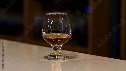 Close up of a glass with cognac, brandy or whisky standing on the bar counter. Stock footage. Strong strong alcoholic beverage inside a glass on the blurred background of a dark room.