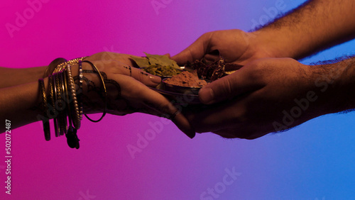 Man passes a plate of spices to indian woman with many bracelets and mehendi on her hands, isolated on bright background. Stock. Man giving set of spices in a plate to woman, indian bazar concept.