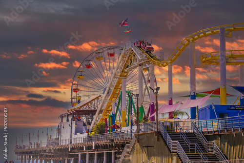 a yellow rollercoaster with flags on top with a colorful Ferris wheel and other colorful carnival games and rides on a brown wooden pier at the beach with powerful clouds at sunset photo