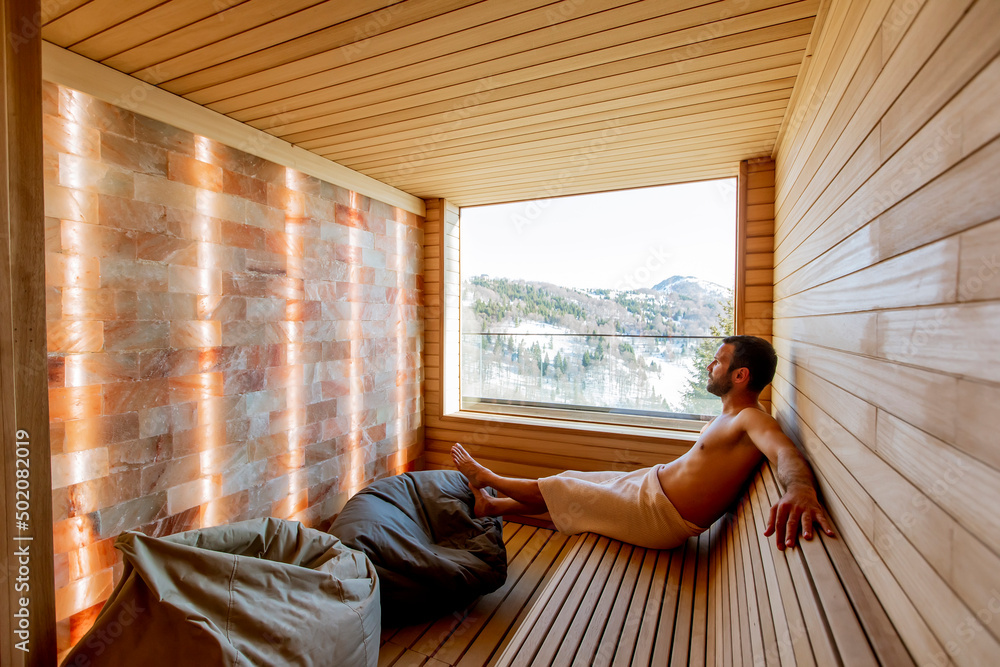 Young man relaxing in the sauna and watching winter forest through the window Young man relaxing in the sauna and watching winter forest through the window