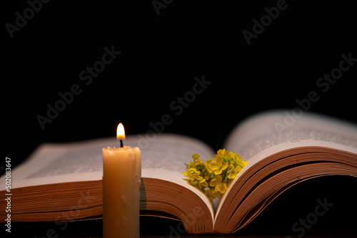 A burning candle, book, and white rose on the table in darkness, space for text. 