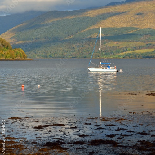 Sailboat anchored in lake. Majestic forest hills. Scotland, UK. Atmospheric landscape. Travel destinations, sailing, cruising, eco tourism, hiking, nature, environmental conservation
