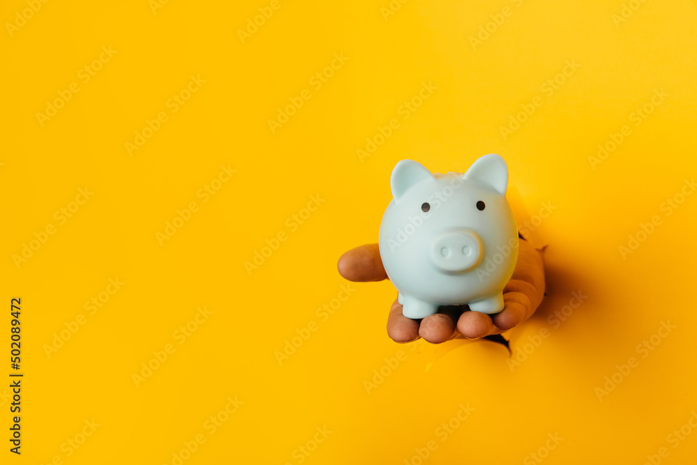 Hand with a blue piggy bank through a yellow paper hole with copy space Hand with a blue piggy bank through a yellow paper hole with copy space