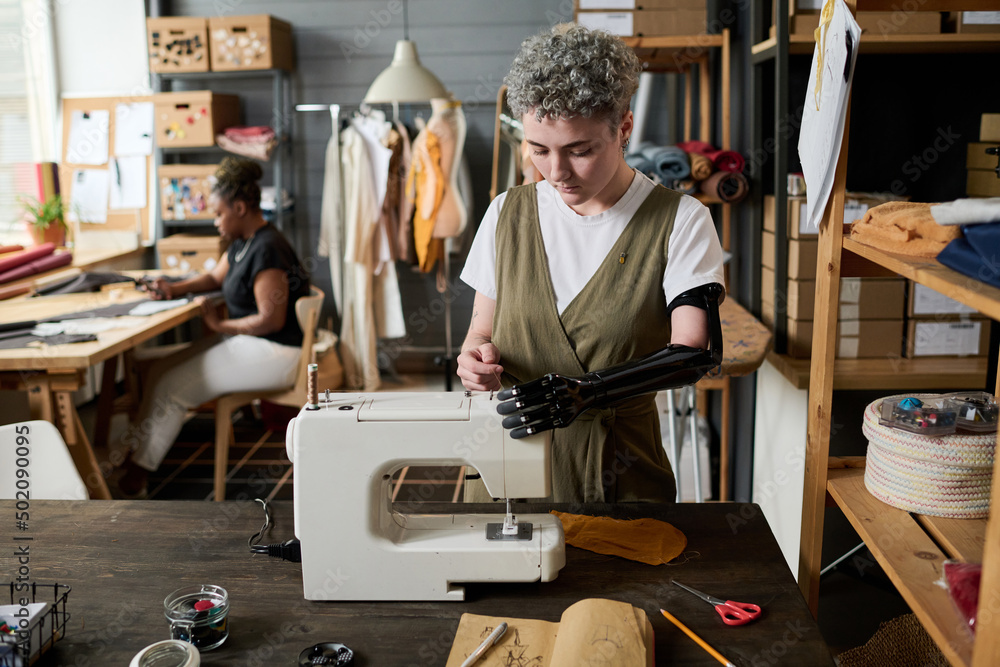 Young female with myoelectric arm fitting thread in sewing machine while standing by her workplace against African American colleague Young female with myoelectric arm fitting thread in sewing machine while standing by her workplace against African American colleague