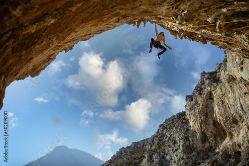 Male rock climber hanging with one hand on on a cliff against sky