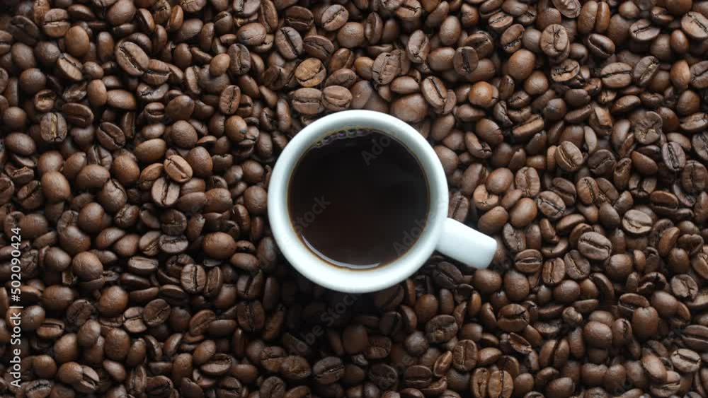 White cup of hot black coffee with a rotating background of fresh roasted coffee beans, close-up, top view White cup of hot black coffee with a rotating background of fresh roasted coffee beans, close-up, top view