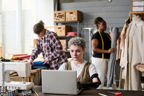 Young serious seamstress or female fashion designer with myoelectric arm working in front of laptop against intercultural colleagues photo