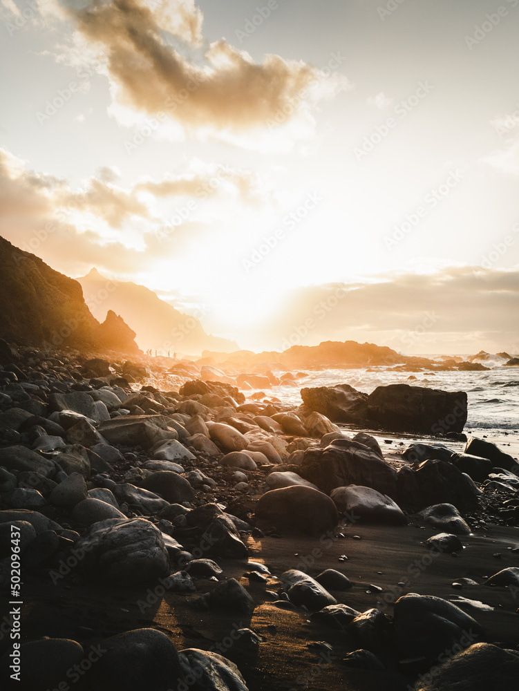 Obraz premium Beautiful view of rocky beach and cliff along North Tenerife coast at sunset. Atlantic wavy ocean with golden sun in Canary Islands.