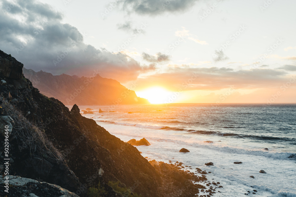 Beautiful scenic view of cliff along North Tenerife coast at sunset. Atlantic wavy ocean with amazing orange sun above the water in Canary Islands.