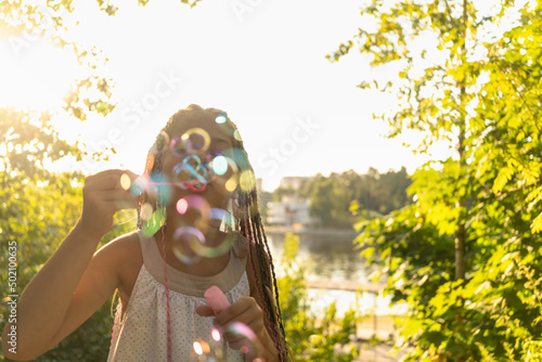 Valokuva cute girl in white dress and with African pigtails blows soap bubbles in park on background of lake