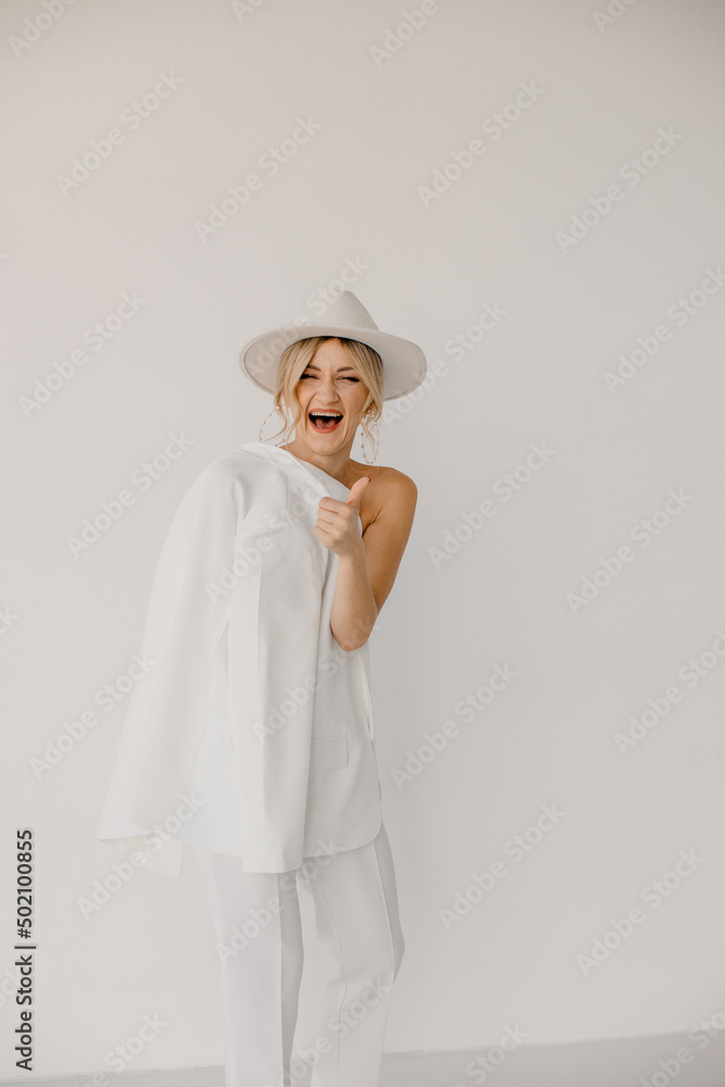 Confident young blond woman smiling, looking at camera isolated on white background. Studio portrait of successful friendly female in white suit and hat, posing over white wall. Confident young blond woman smiling, looking at camera isolated on white background. Studio portrait of successful friendly female in white suit and hat, posing over white wall.