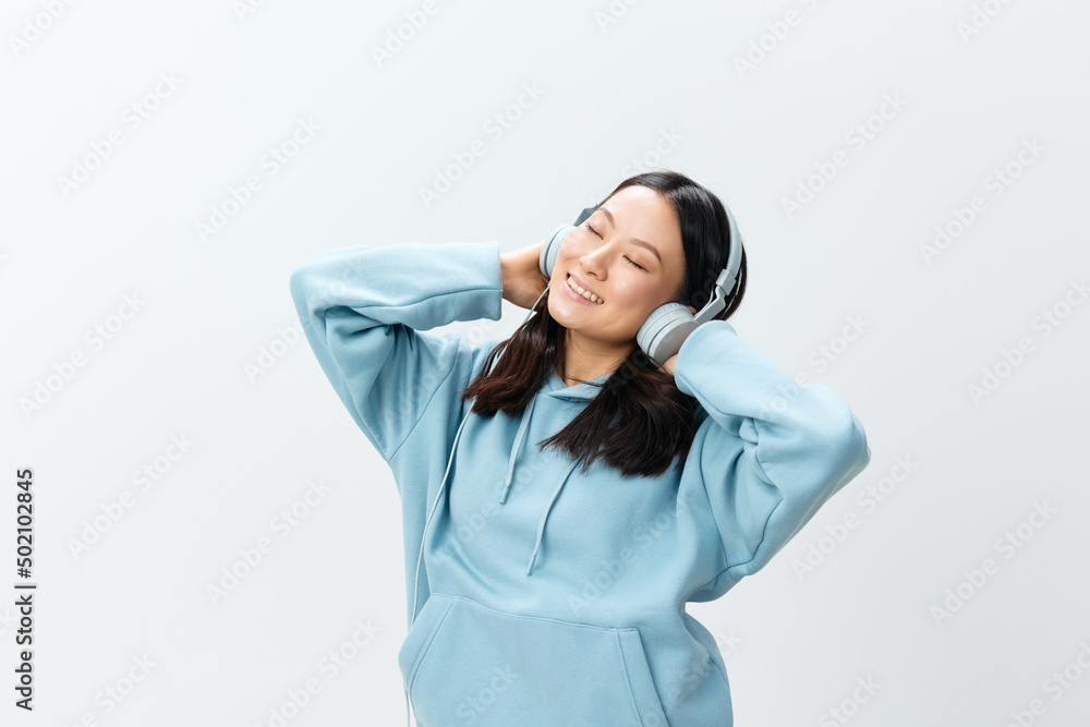 Overjoyed happy pretty Korean young female in blue hoodie touch headphones relax closing eyes posing isolated on over white studio background. The best offer for ad. Music App Platform Ad concept Overjoyed happy pretty Korean young female in blue hoodie touch headphones relax closing eyes posing isolated on over white studio background. The best offer for ad. Music App Platform Ad concept
