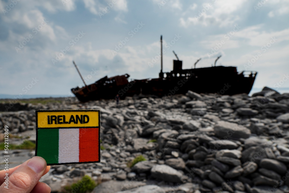Irish flag and sign Ireland in focus. Plassey shipwreck on shore of Inisheer island. Aran islands, county Galway, Ireland out of focus in the background. Travel and tourism industry. Irish flag and sign Ireland in focus. Plassey shipwreck on shore of Inisheer island. Aran islands, county Galway, Ireland out of focus in the background. Travel and tourism industry.