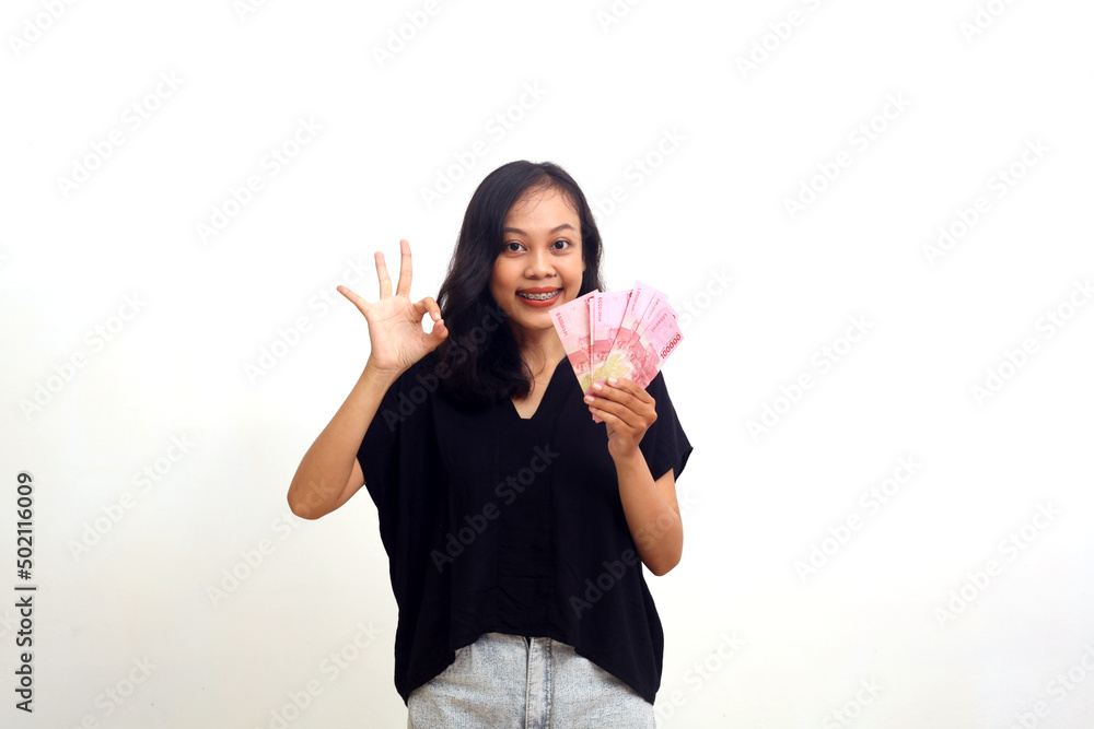 Happy asian young woman standing while holding indonesian money and giving an okay gesture. Happy asian young woman standing while holding indonesian money and giving an okay gesture.