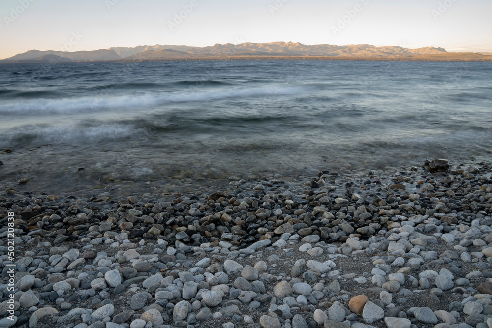 Long exposure shot of Nahuel Huapi lake at sunset. Beautiful blurred water effect, the rocky shore and Andes mountains in the horizon with a magical dusk light. Long exposure shot of Nahuel Huapi lake at sunset. Beautiful blurred water effect, the rocky shore and Andes mountains in the horizon with a magical dusk light.
