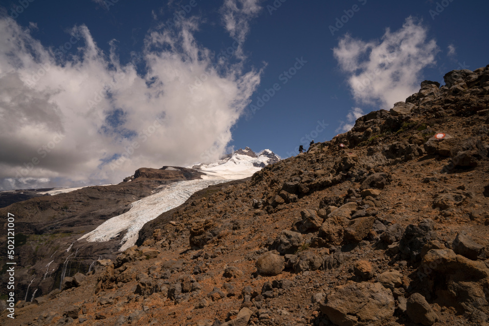 Alpine scenic. View of two hikers climbing up Tronador hill and glacier Castaño Overo in the Andes mountains in Patagonia Argentina. The rocky mountaintop and glacier ice field under a majestic sky.