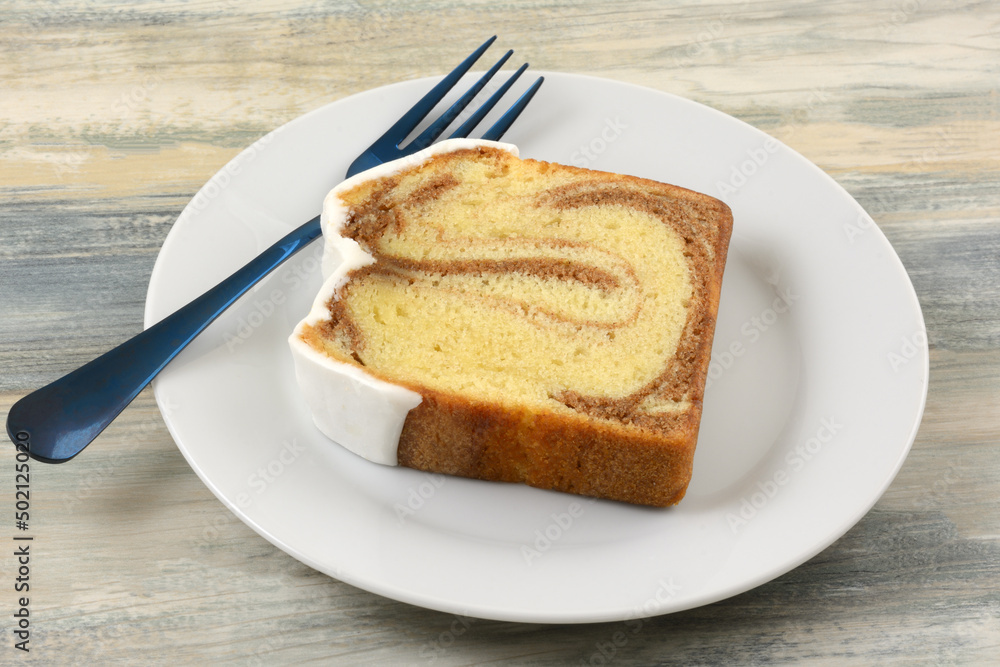 Slice of cinnamon swirl cake with icing on white plate with blue fork