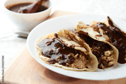 Tacos of mole with rice, placed on wood, in the background a piece of chicken